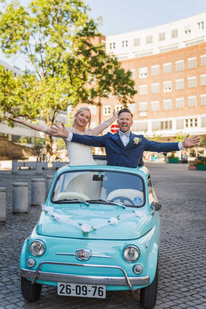 Brudepar som står i en liten Fiat med takluke og strekker armene i været på en sidegate ved Oslo Rådhus, et lekent bryllupsbilde.