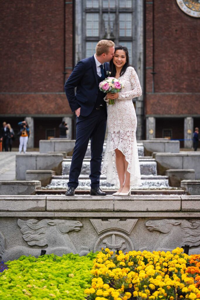 Brudepar ved vanntrappen utenfor Oslo Rådhus med gule blomster i forgrunnen, der brudgommen kysser bruden på kinnet mens hun ser inn i kameraet.
