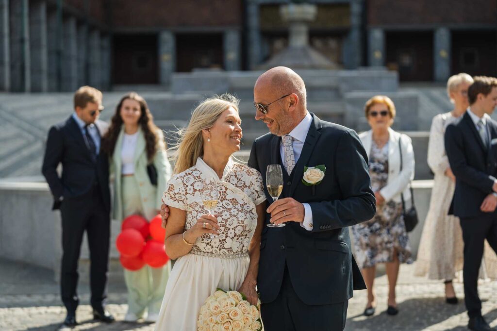 Brudepar som skåler i champagne i Borggården foran Oslo Rådhus, med gjester og den ikoniske arkitekturen som stemningsfull bakgrunn.
