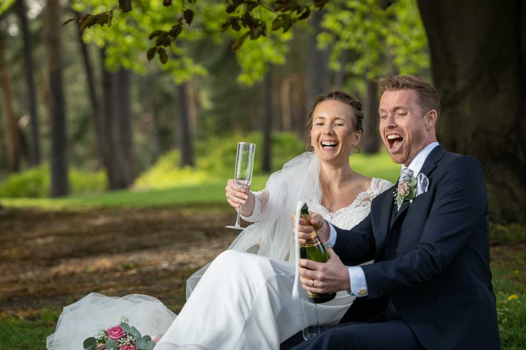 Brudeparet sitter i gresset og åpner en flaske champagne som spruter av skum.