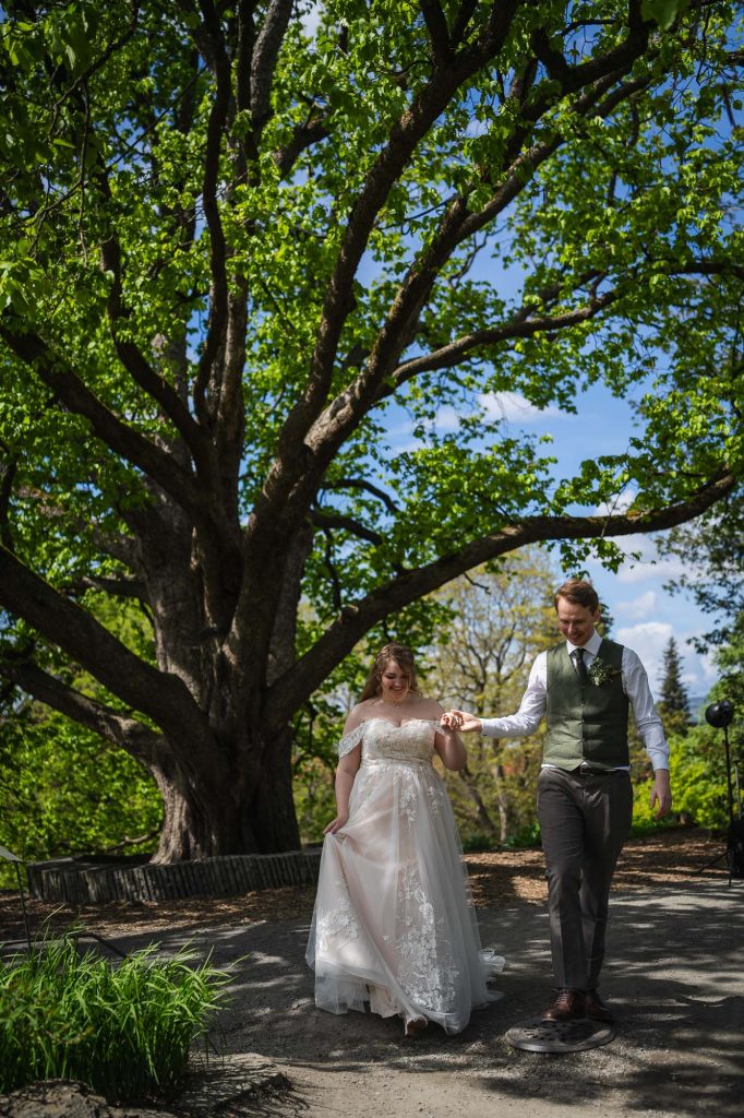 Brudeparet går hånd i hånd foran et stort tre i botanisk hage i Oslo.