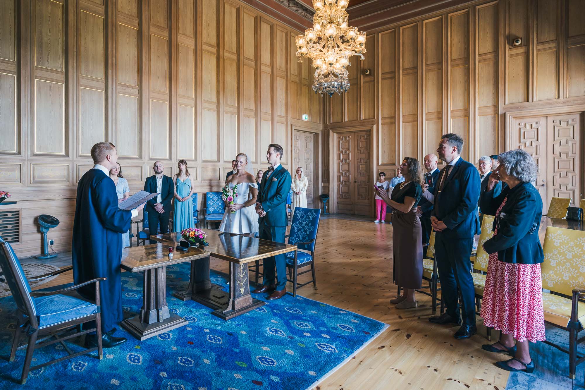 Et brudepar under en intim vielse i Munch-rommet i Oslo rådhus, omgitt av sine nærmeste gjester foran de berømte veggmaleriene.