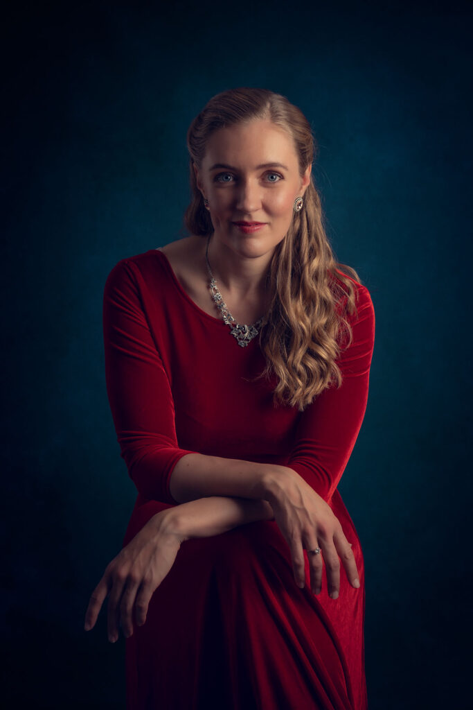 Et elegant studioportrett av en kvinne i rød fløyelskjole mot en dyp blå bakgrunn, fotografert med klassisk lyssetting.