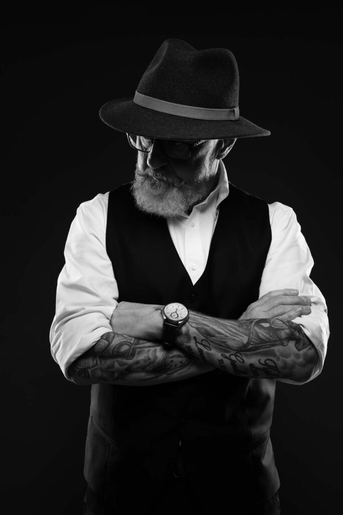 Et tøft og karakterfullt sort-hvitt-portrett av en eldre mann med hatt, skjegg og tatoverte armer i kors, fotografert i studio.