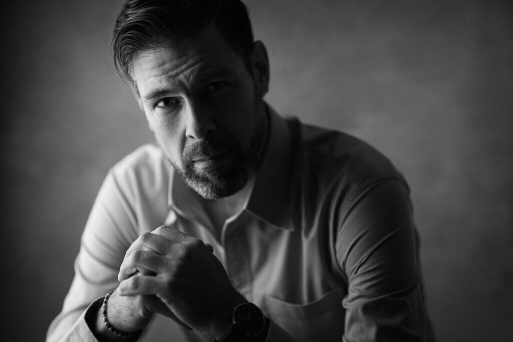 Et klassisk sort-hvitt-portrett av en mann i skjorte som hviler haken mot knyttede hender, fotografert med fokus på et intenst blikk.