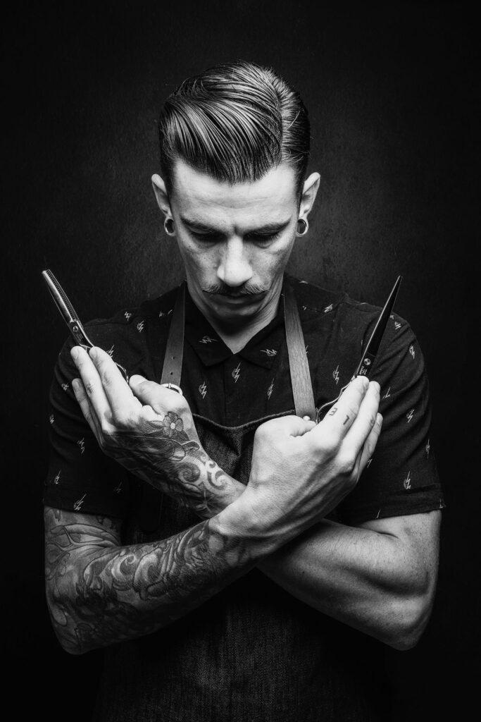 Stemningsfullt karakterportrett av en mann med tatoveringer og sakser, fotografert med dramatisk lys i studio.