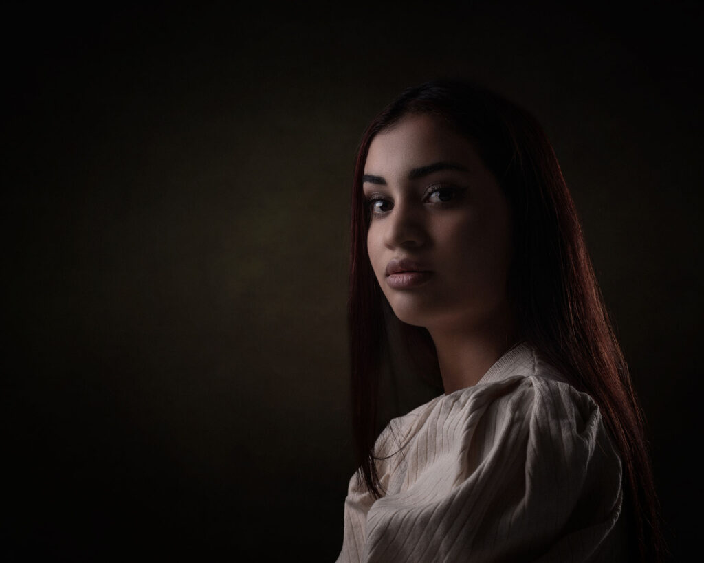 Et stemningsfullt portrett av en ung kvinne med langt mørkt hår, fotografert i studio med myk lyssetting mot en mørk bakgrunn.