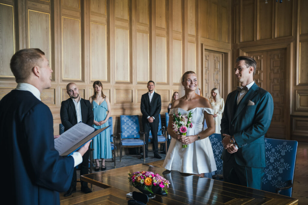 Et lykkelig og smilende brudepar under den borgerlige vielsen i Munchrommet på Oslo rådhus, med gjester i bakgrunnen.